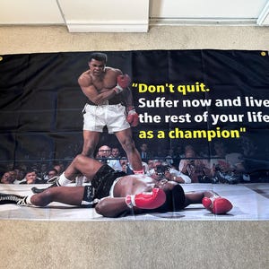 Könnte beinhalten: Schwarzes Banner mit einer Boxszene mit Muhammad Ali. Auf dem Banner steht das Zitat: „Gib nicht auf. Leide jetzt und lebe den Rest deines Lebens als Champion.“ Das Bild zeigt einen Boxer, der über seinem Gegner steht.