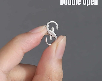 Può includere: Un anello in metallo color argento con design a doppia apertura, caratterizzato da una finitura liscia e lucida. L'anello presenta una struttura curva e sovrapposta che crea una forma a S. Il testo Double Open è visibile nell'angolo in alto a destra.