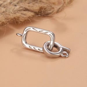 May include: A silver-toned metal jewellery clasp consisting of a rectangular link with etched diagonal lines and a circular spring ring attached to one side. The rectangular component features a small loop on the opposite end for secure attachment.