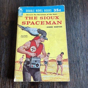 May include: Vintage paperback book titled "The Sioux Spaceman" by Andre Norton. The cover features a retro illustration of a spaceman with a bird-like mask and a red vest, holding a radio. The book is a "Double Novel Book" and is marked as a "First Book Publication".