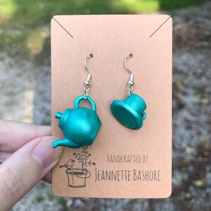 May include: Teal-colored teapot and hat earrings with silver hooks. The earrings are displayed on a brown card with the text "Handcrafted by Jeannette Bashore" and a small illustration of a bird and a bucket.