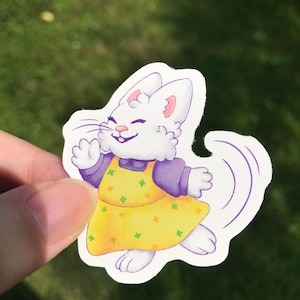 May include: A sticker featuring a cheerful white rabbit with a smiling face. The rabbit wears a yellow dress with green and orange floral accents and purple sleeves. The sticker has a white border and is held against a green background.