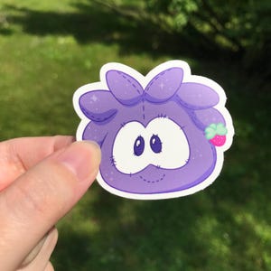 May include: A purple sticker featuring a cartoon character with large eyes and a stitched design. The character has a small strawberry accessory. The sticker is held in a hand against a blurred green background.