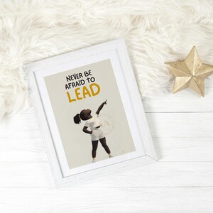 May include: White framed art print with the text "NEVER BE AFRAID TO LEAD" and an illustration of a person pointing upwards. A gold star decoration is next to the frame on a white wooden surface.