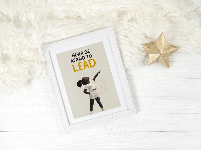 May include: White framed art print with the text "NEVER BE AFRAID TO LEAD" and an illustration of a person pointing upwards. A gold star decoration is next to the frame on a white wooden surface.