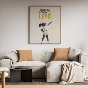 May include: A modern living room with a light gray sectional sofa, two brown throw pillows, and a framed art print that reads "NEVER BE AFRAID TO LEAD". The room includes a wooden bookshelf, a side table, and a chair.