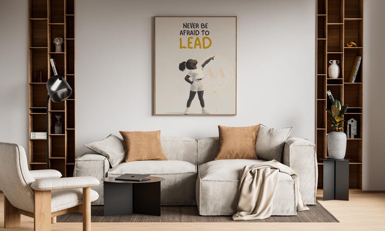 May include: A modern living room with a light gray sectional sofa, two brown throw pillows, and a framed art print that reads "NEVER BE AFRAID TO LEAD". The room includes a wooden bookshelf, a side table, and a chair.