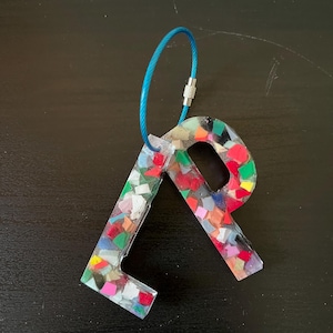 May include: A colorful letter "R" keychain made from recycled materials. The keychain features a mix of red, green, blue, and white fragments encased in clear resin. A blue wire loop is attached for easy use.