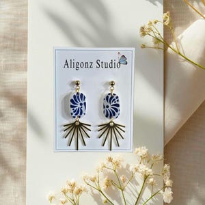 May include: A pair of earrings with a blue and white floral design on an oval shape, topped with a gold stud and connected to a gold sunburst design. The earrings are displayed on a white card with the text "Aligonz Studio".