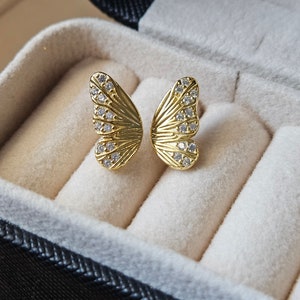 May include: A pair of gold-colored butterfly wing earrings with small, clear gemstones. The earrings are displayed in a black jewelry box with a beige interior. The butterfly wings have detailed vein patterns.