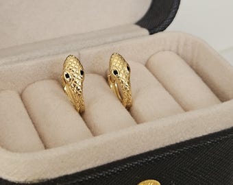 May include: A pair of gold snake hoop earrings with black eyes, displayed in a black velvet jewelry box.