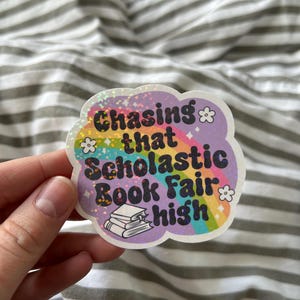 May include: A purple and rainbow sticker with the text "Chasing that Scholastic Book Fair high" and a stack of books. The sticker has a glittery finish.