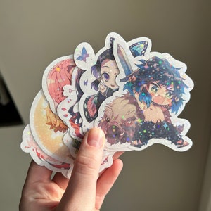 Peut inclure: Un ensemble de quatre autocollants holographiques de personnages d'anime mettant en scène des personnages de la série d'anime populaire Demon Slayer. Les autocollants représentent Inosuke, Zenitsu, Tanjiro et Nezuko. Les autocollants sont imprimés sur du vinyle transparent et ont une finition holographique.