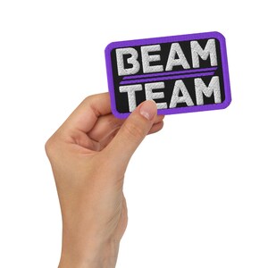 Puede incluir: Un parche bordado de color morado y negro con el texto "BEAM TEAM" en blanco.