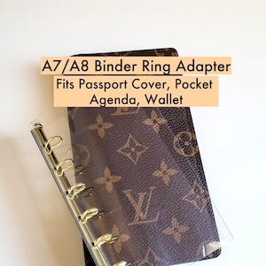 Puede incluir: Un adaptador de anilla de encuadernación A7/A8 transparente con anillas doradas, mostrado sobre una cartera estampada marrón y beige. El texto de la imagen dice: A7/A8 Binder Ring Adapter Fits Passport Cover, Pocket Agenda, Wallet.
