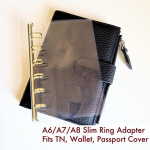 May include: A clear plastic ring adapter with a gold-colored metal spine featuring six rings, placed over a black textured wallet. The text A6/A7/A8 Slim Ring Adapter Fits TN, Wallet, Passport Cover is printed in dark red at the bottom of the image.