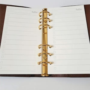 May include: Open planner with lined pages and a gold-colored binder mechanism. The pages are white with the word "To Do" at the top. The binder is a shiny gold color, and the planner has a brown cover.