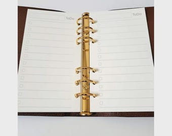 May include: Open planner with lined pages and a gold-colored binder mechanism. The pages are white with the word "To Do" at the top. The binder is a shiny gold color, and the planner has a brown cover.