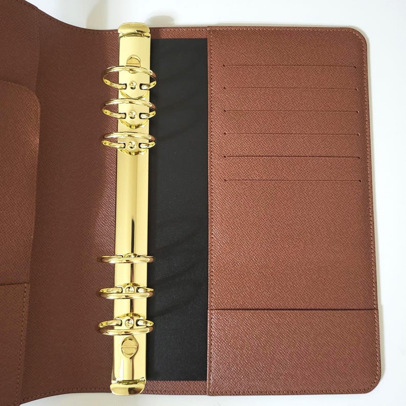 May include: A brown textured leather planner binder featuring a gold-toned six-ring metal mechanism. The interior includes multiple horizontal card slots on the right side and a vertical pocket on the left, with a black spine accent.