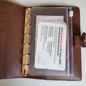 May include: A brown textured leather planner with a gold six-ring binder mechanism. Inside, a clear plastic zippered pouch holds a white Caremark insurance card with text including Visit Caremark.com for easy refills and more.