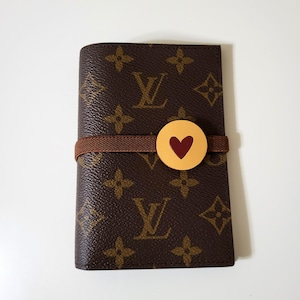 May include: Brown leather wallet with a repeating logo pattern and a brown elastic band. A gold-colored circular accent with a red heart is attached to the band. The wallet is closed.