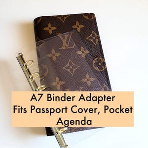 May include: A brown A7 binder adapter with gold hardware, designed to fit passport covers, pocket agendas, and other small items. The text on the image reads: "A7 Binder Adapter Fits Passport Cover, Pocket Agenda."