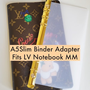 May include: A brown notebook with a gold binder and a clear plastic sheet. The notebook has a monogram pattern and the text "A5Slim Binder Adapter Fits LV Notebook MM". An embroidered green and blue design is visible.