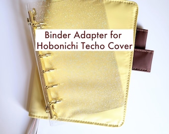 May include: A clear, glitter-infused plastic binder adapter with gold-toned metal rings, positioned over a pale yellow notebook cover. The text "Binder Adapter for Hobonichi Techo Cover" is printed in dark red across the center of the image.