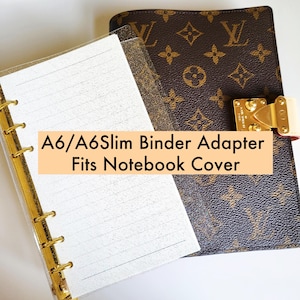 May include: An A6/A6Slim binder adapter fits a notebook cover. The notebook cover is brown with a repeating pattern and a gold clasp. The adapter has a gold spine and lined white paper.