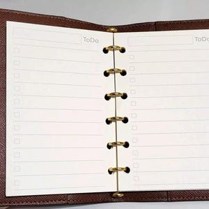 May include: Open brown leather planner with lined To Do list pages. The pages are held together by gold-colored rings. The top of each page has the word "To Do".