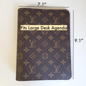 May include: A brown and tan monogram patterned agenda cover featuring the text Fits Large Desk Agenda. The cover measures 7.1 inches in width and 9.1 inches in height, displaying a repeating logo design across the entire front surface.