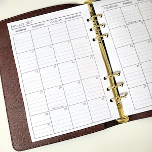 May include: A brown leather-textured planner open to a January 2027 monthly calendar page. The white paper features a grid layout with black text for days of the week and dates, secured by a gold-colored six-ring metal binder mechanism.