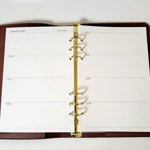 May include: An open brown leather planner with a gold-toned six-ring metal binder mechanism. The white pages feature a weekly plan layout with headings for Monday, Tuesday, Wednesday, Thursday, Friday, Saturday, and Sunday printed in black ink.
