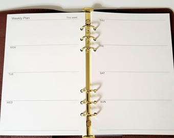 May include: An open brown leather planner with a gold-toned six-ring metal binder mechanism. The white pages feature a weekly plan layout with headings for Monday, Tuesday, Wednesday, Thursday, Friday, Saturday, and Sunday printed in black ink.