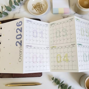 May include: An open planner with a 2026 calendar overview. The calendar pages are white with pastel-colored highlighting and text. A gold pen, coffee cup, and small dish with paper clips are also visible.