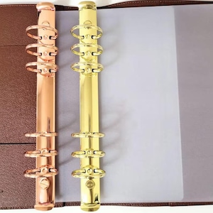 May include: A brown textured binder featuring two vertical metal ring mechanisms. One mechanism is rose gold and the other is gold, each containing six circular rings for holding paper. The binder includes a translucent plastic sheet.