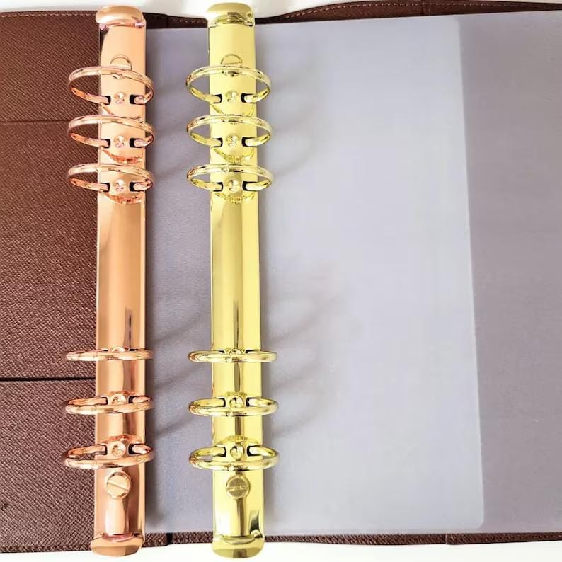 May include: A brown textured binder featuring two vertical metal ring mechanisms. One mechanism is rose gold and the other is gold, each containing six circular rings for holding paper. The binder includes a translucent plastic sheet.