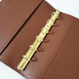May include: Open brown leather binder with a gold-colored, six-ring binder mechanism. The binder is open, revealing the interior with card slots. The gold rings are aligned vertically, and the leather has a textured appearance.