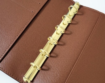 May include: Open brown leather binder with a gold-colored, six-ring binder mechanism. The binder is open, revealing the interior with card slots. The gold rings are aligned vertically, and the leather has a textured appearance.