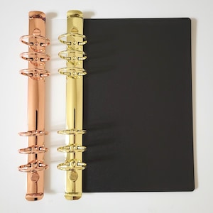 May include: Two metal binder mechanisms with six rings each, one in a polished rose gold finish and one in a bright gold finish, attached to a rectangular black cover. The mechanisms feature a vertical spine with three rings at the top and three at the bottom.
