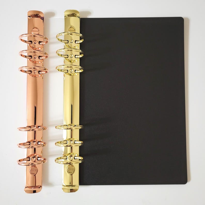 May include: Two metal binder mechanisms with six rings each, one in a polished rose gold finish and one in a bright gold finish, attached to a rectangular black cover. The mechanisms feature a vertical spine with three rings at the top and three at the bottom.
