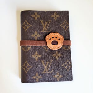 May include: A brown passport holder featuring a repeating dark brown and tan monogram pattern. A brown elastic band wraps horizontally around the center, secured by a light tan leather patch shaped like a paw print with four small circular cutouts.