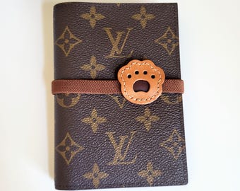 May include: A brown passport holder featuring a repeating dark brown and tan monogram pattern. A brown elastic band wraps horizontally around the center, secured by a light tan leather patch shaped like a paw print with four small circular cutouts.
