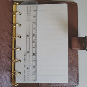 May include: A brown leather planner with a gold six-ring binder mechanism. A clear plastic ruler is placed over the white lined paper, showing measurements up to 4 inches. The ruler features the text TODAY and FOR DAILY USE.