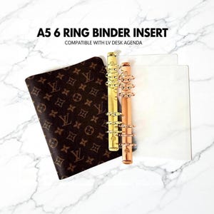 May include: A5 6 ring binder insert compatible with LV desk agenda. The set includes a brown cover with a repeating monogram pattern, a gold-toned metal six-ring mechanism, a rose gold-toned metal six-ring mechanism, and a clear plastic sheet.