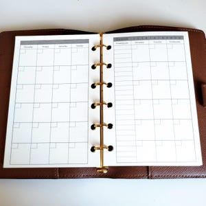 May include: Open planner with a brown leather cover and gold-colored metal rings. The planner is open to a monthly calendar page with a grid layout. The calendar page is white with black lines and text.