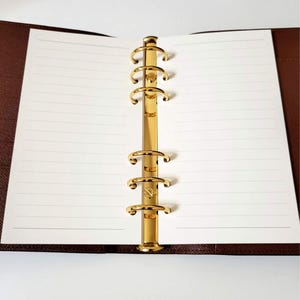 May include: A brown leather bound notebook with gold rings and lined white paper.