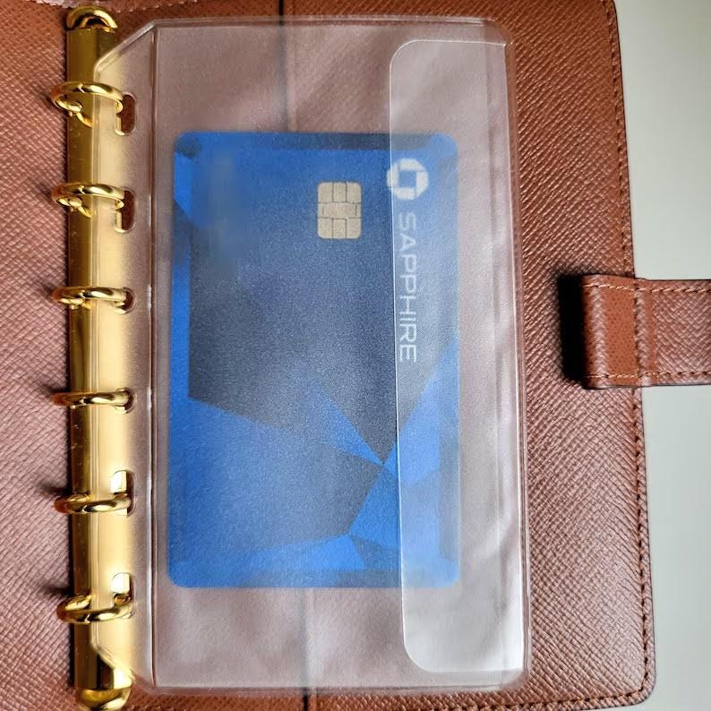 May include: A brown leather planner with a gold-toned six-ring metal binder mechanism. A clear plastic pocket is attached to the rings, holding a blue credit card with the text SAPPHIRE and a gold chip visible on the front surface.
