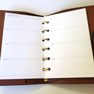 May include: A brown leather bound weekly planner with gold rings and a white paper insert. The paper insert has the days of the week listed: Mon, Tue, Wed, Fri, Sat, Sun.