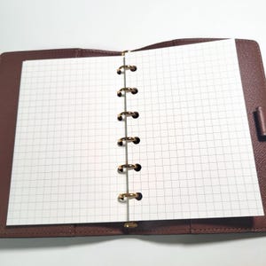 May include: Open brown leather notebook with a grid-lined paper insert. The notebook is open, revealing the blank pages. The binding features gold-colored rings. The notebook is on a white surface.